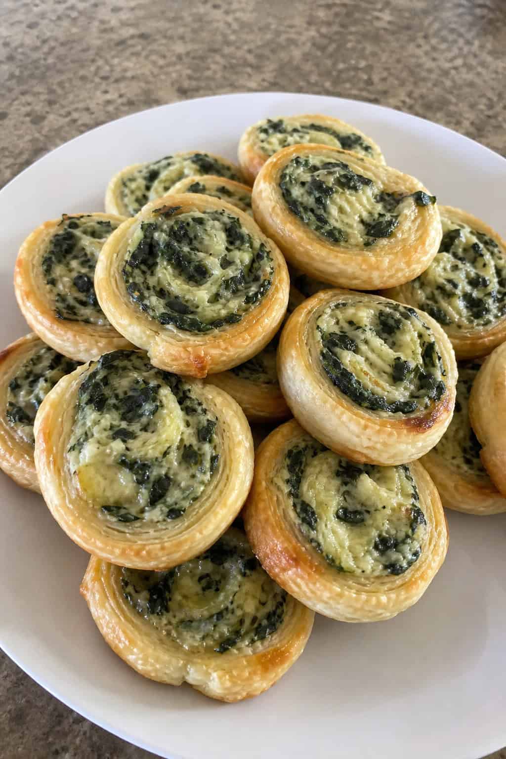 Close-up of spinach artichoke dip pinwheels on a wooden board showing golden-edged puff pastry swirls filled with creamy green and white filling, with a bowl of dip and game day items visible in the background.