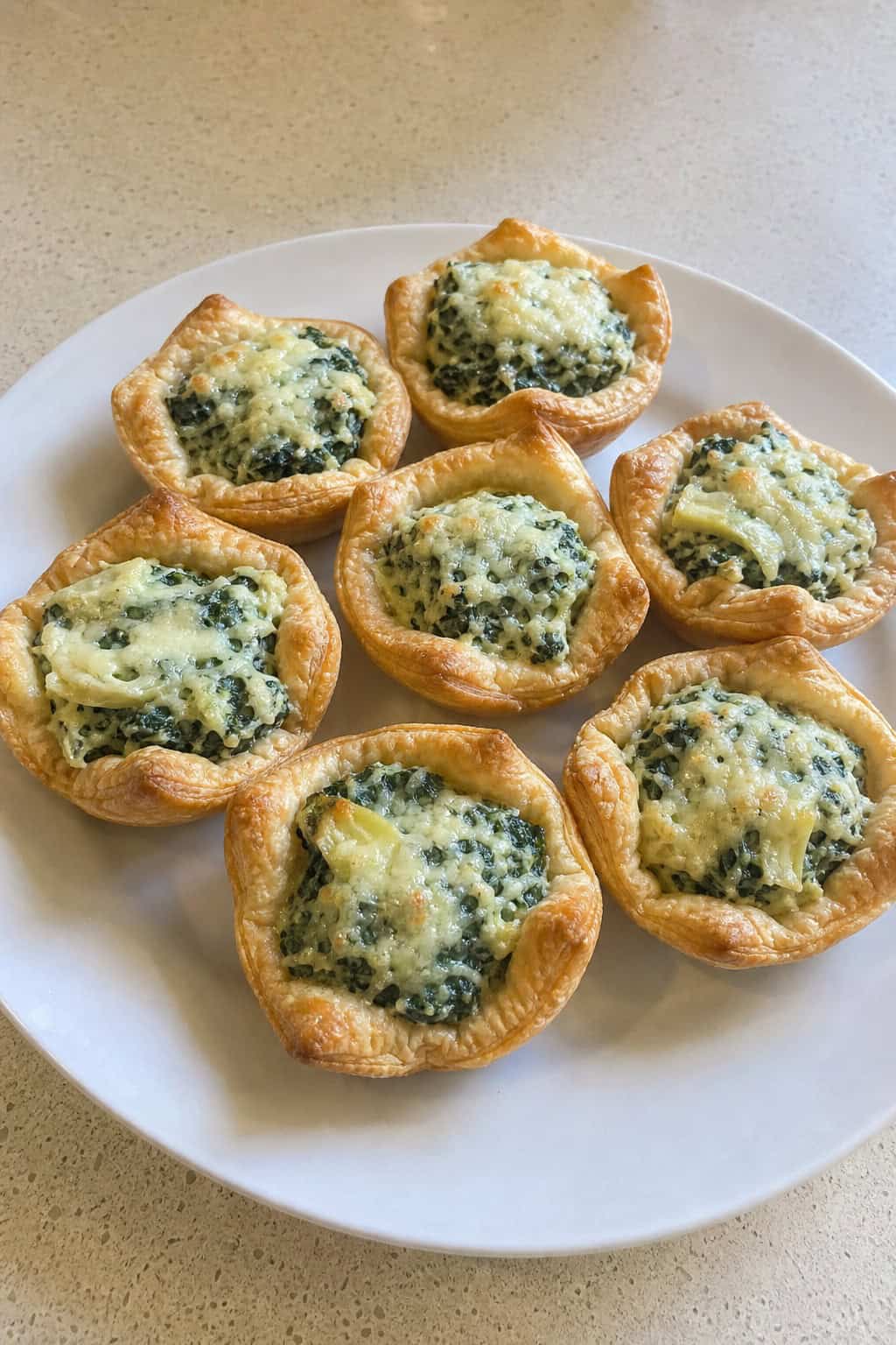 Close-up of golden puff pastry cups filled with creamy spinach and artichoke dip, arranged on a wooden board with fresh spinach leaves nearby.