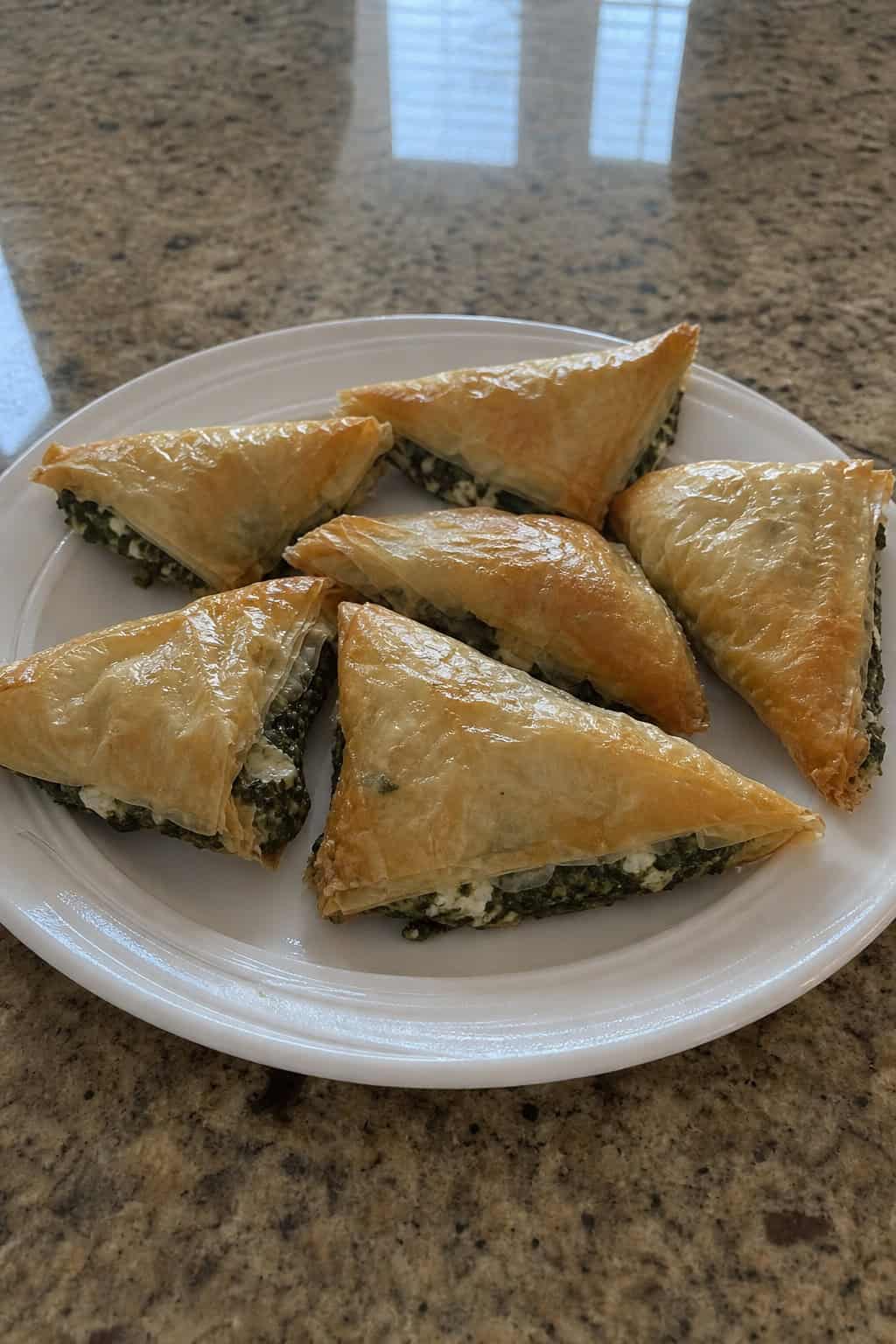Close-up of golden spinach and feta phyllo triangles arranged on a wooden cutting board with fresh spinach and feta cheese in the background.