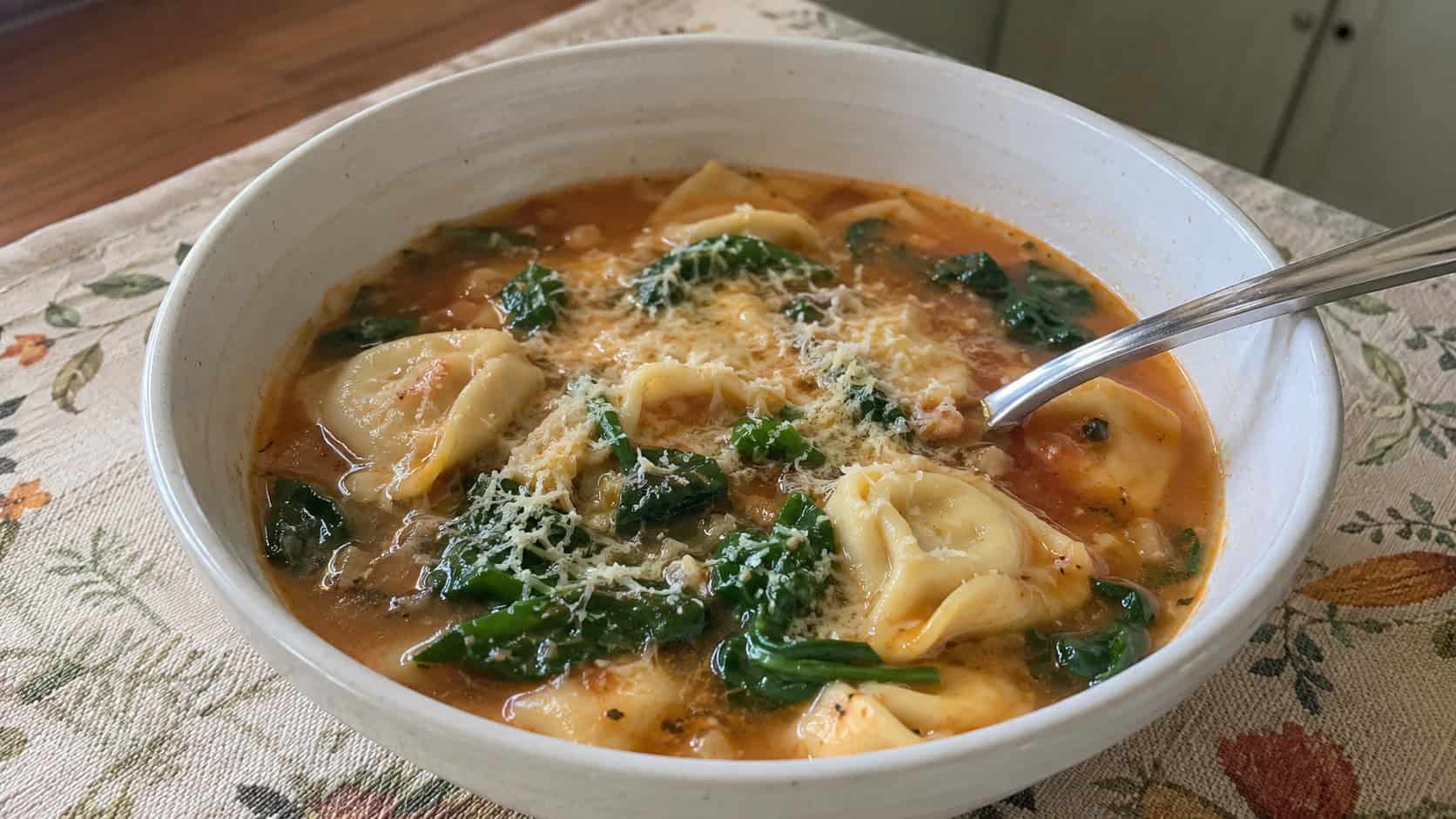 a finished bowl of spinach tortellini soup in a light tomato broth, plump cheese tortellini and bright wilted spinach visible, grated Parmesan on top, spoon in the bowl, warm natural kitchen light, casual home cooking