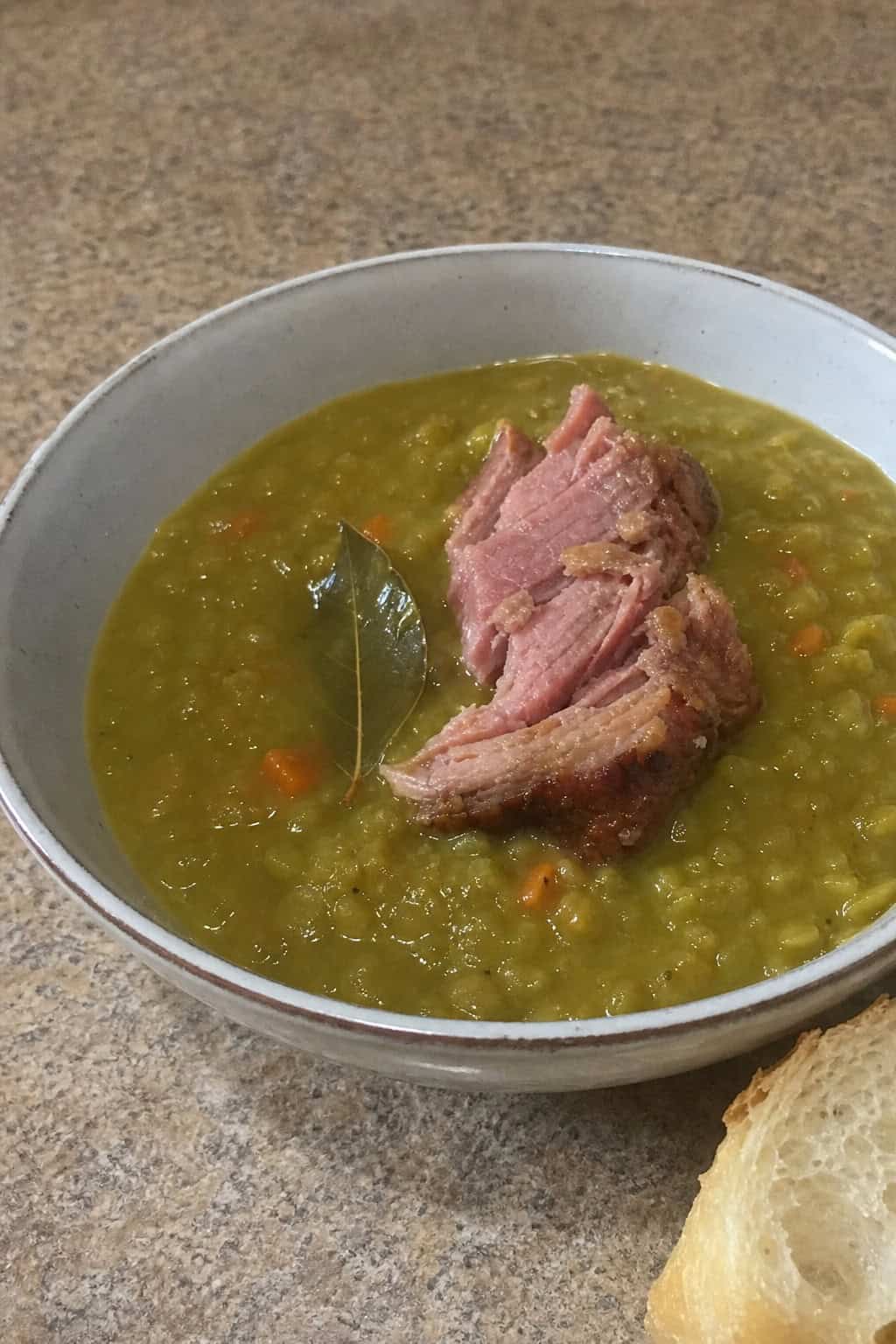 A bowl of split pea soup with a ham hock and bay leaf on a wooden table surrounded by bread and herbs.