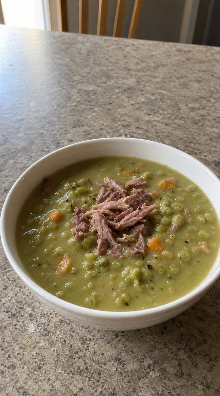 A bowl of green split pea soup with smoked ham hock on a wooden table, surrounded by fresh ingredients.
