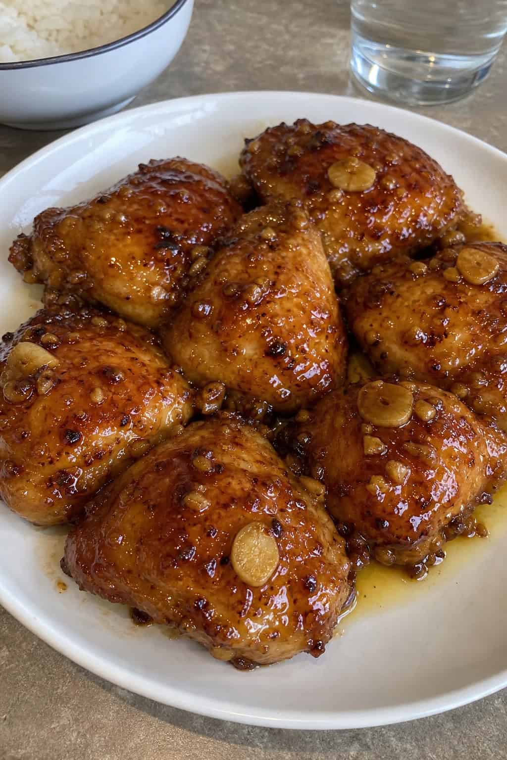 A plate of sticky honey garlic baked chicken thighs garnished with parsley and garlic slices on a wooden table.