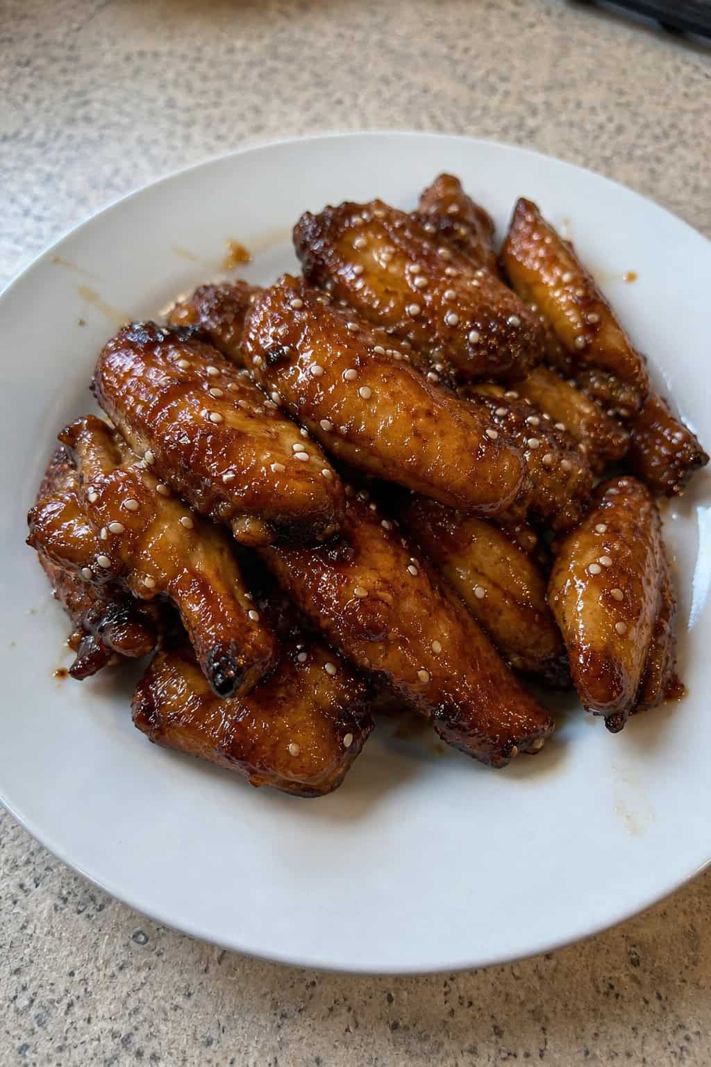 A plate of sticky honey garlic chicken wings garnished with sesame seeds and green onions, with a bowl of soy sauce and chopsticks on a wooden table.