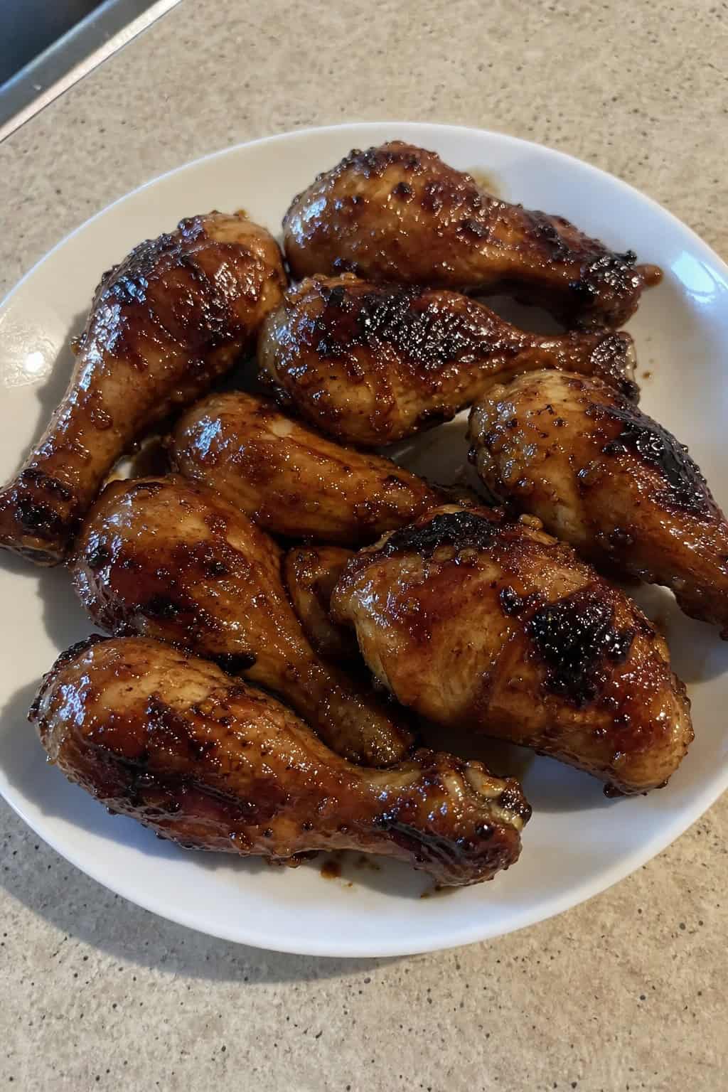A white plate of sticky baked chicken drumsticks with a dark caramelized maple-soy glaze, topped with thinly sliced green onions and a light sprinkle of sesame seeds.