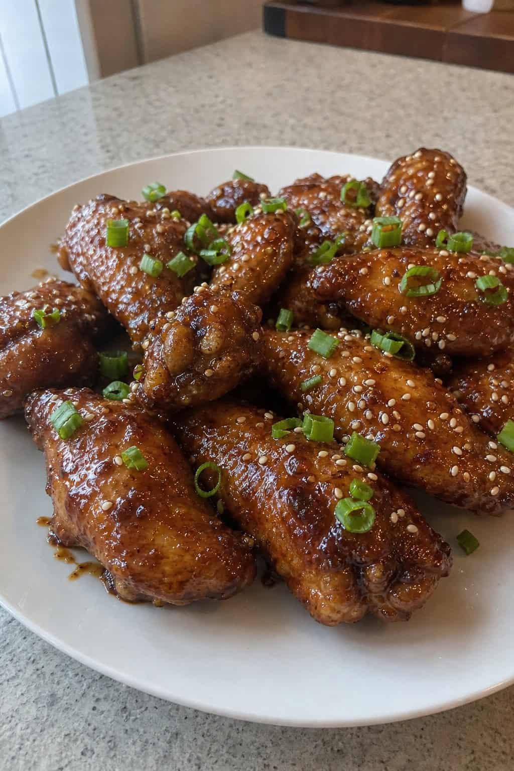 A plate of sticky teriyaki chicken wings garnished with green onions and sesame seeds on a wooden board, with blurred crock pot and dipping sauces in the background.