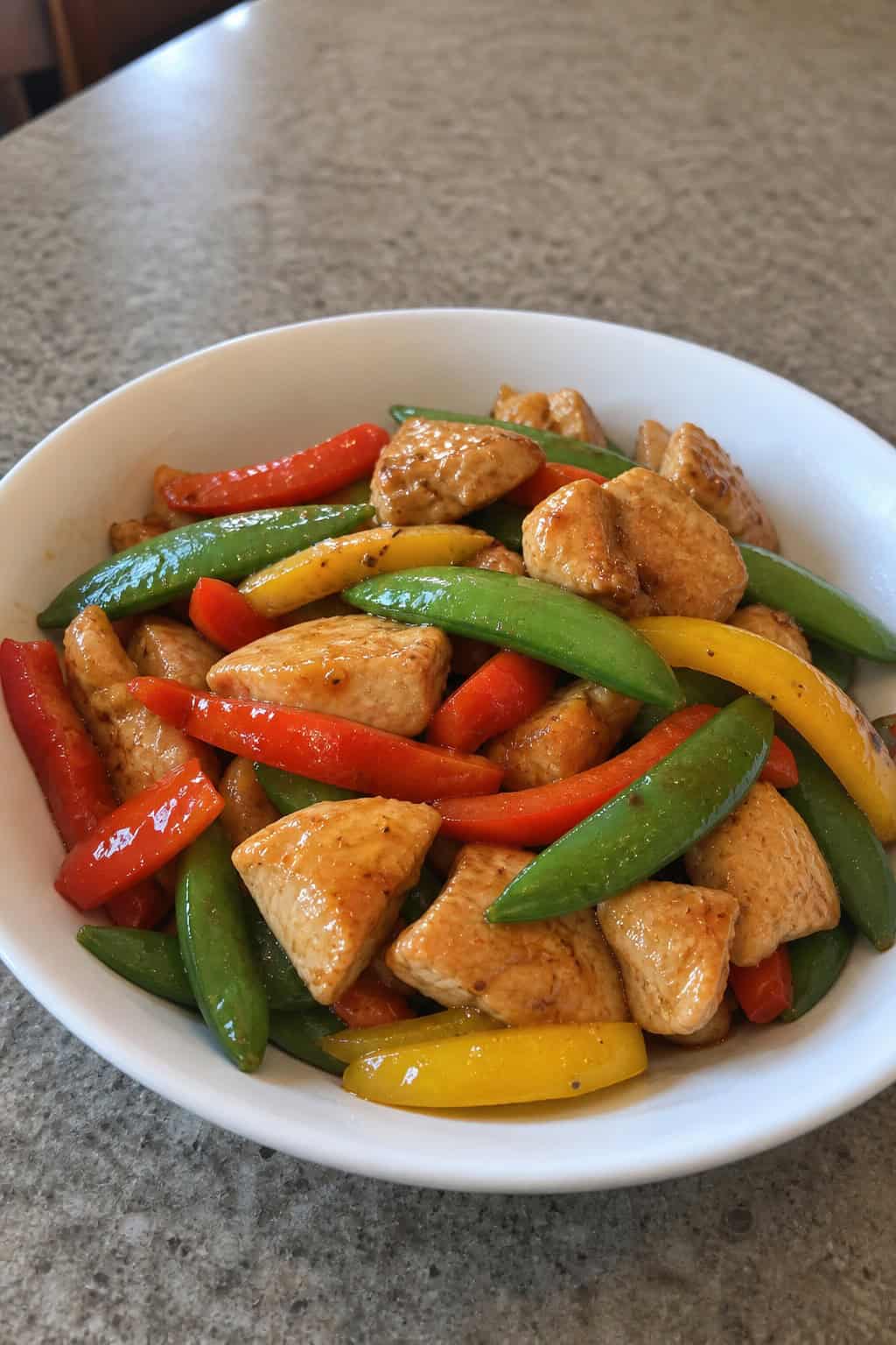 A bowl of stir-fry chicken with snap peas and red and yellow bell peppers on a wooden table.