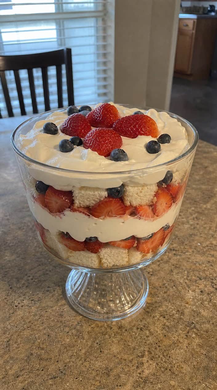 A layered dessert of angel food cake, strawberries, and whipped cream in a clear glass bowl with patriotic decorations in the background.