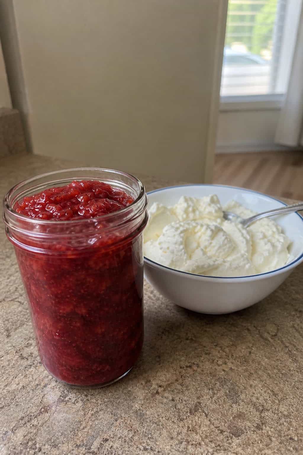 A jar of strawberry jam and a bowl of creamy cheesecake filling on a wooden countertop with fresh strawberries around them.