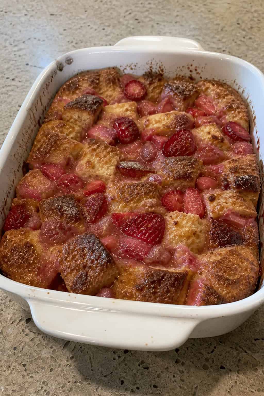 A baked strawberry-rhubarb French toast casserole in a deep ceramic dish with a golden-brown top, surrounded by fresh strawberries and cut rhubarb stalks on a wooden table.