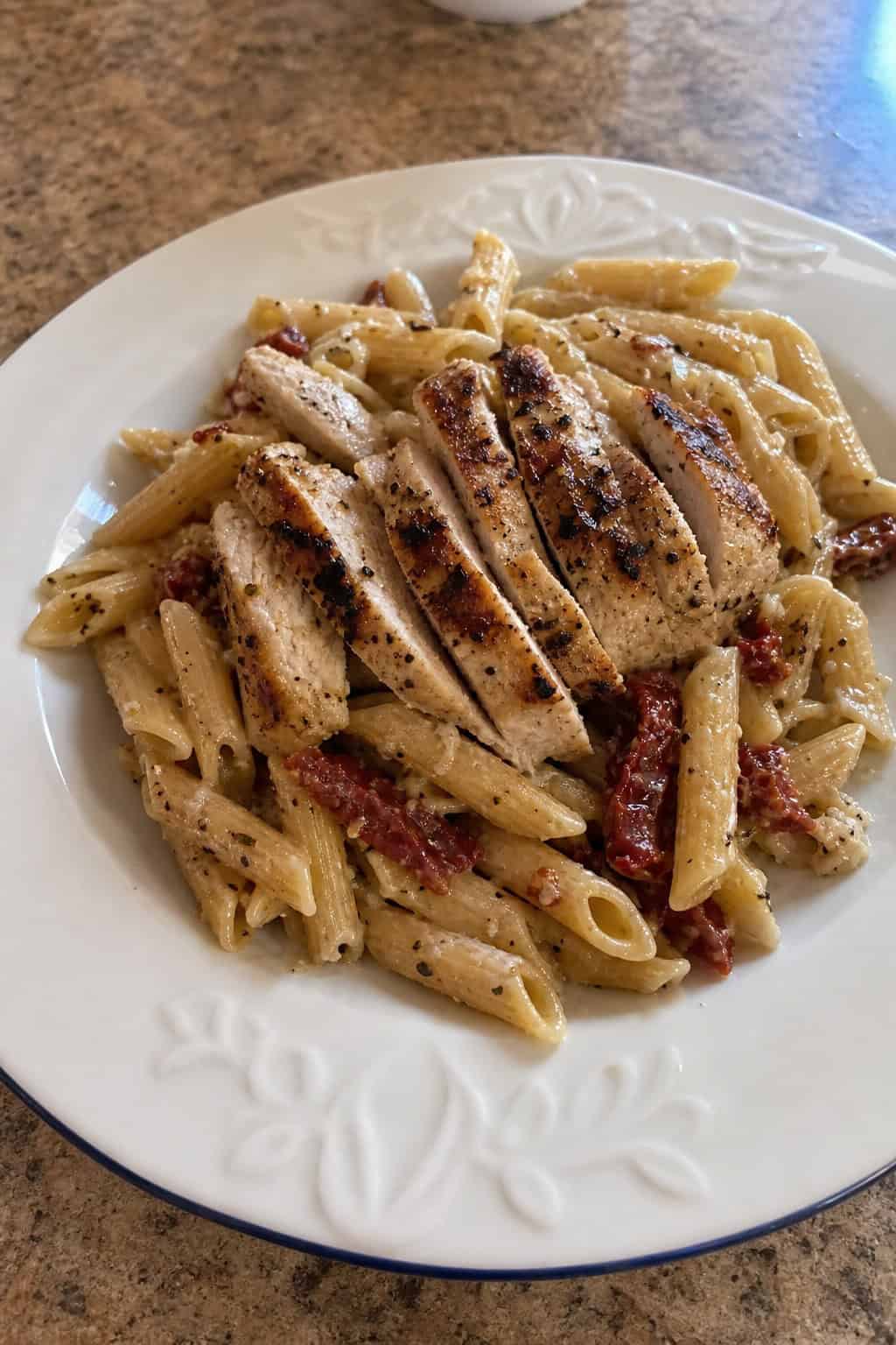 Plate of sun-dried tomato chicken pasta with grilled chicken, sun-dried tomatoes, basil, and Parmesan cheese on a wooden table.