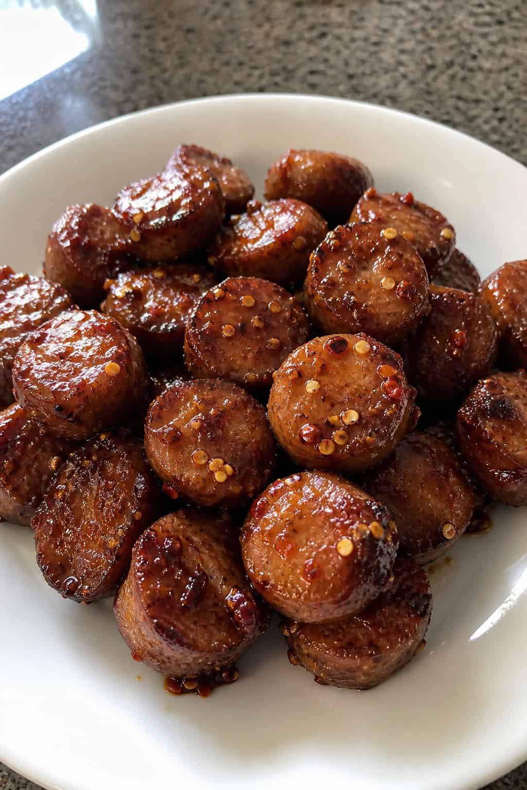 Close-up of glazed sausage bites sprinkled with red pepper flakes on a wooden serving platter, garnished with fresh herbs.