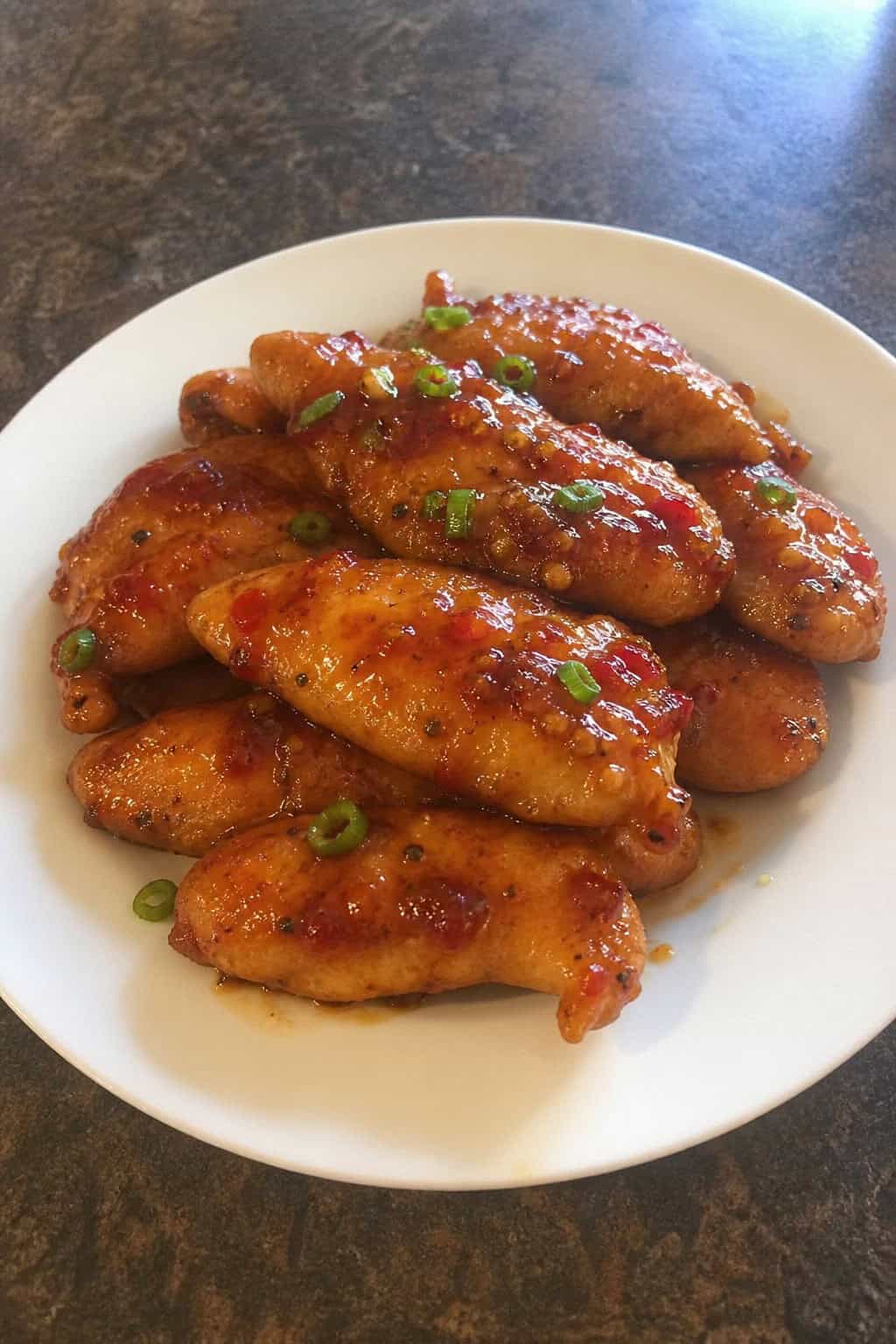 Glossy sweet chili glazed chicken tenderloins garnished with sliced green onions on a wooden table, with fresh chili peppers and lime wedges arranged nearby.