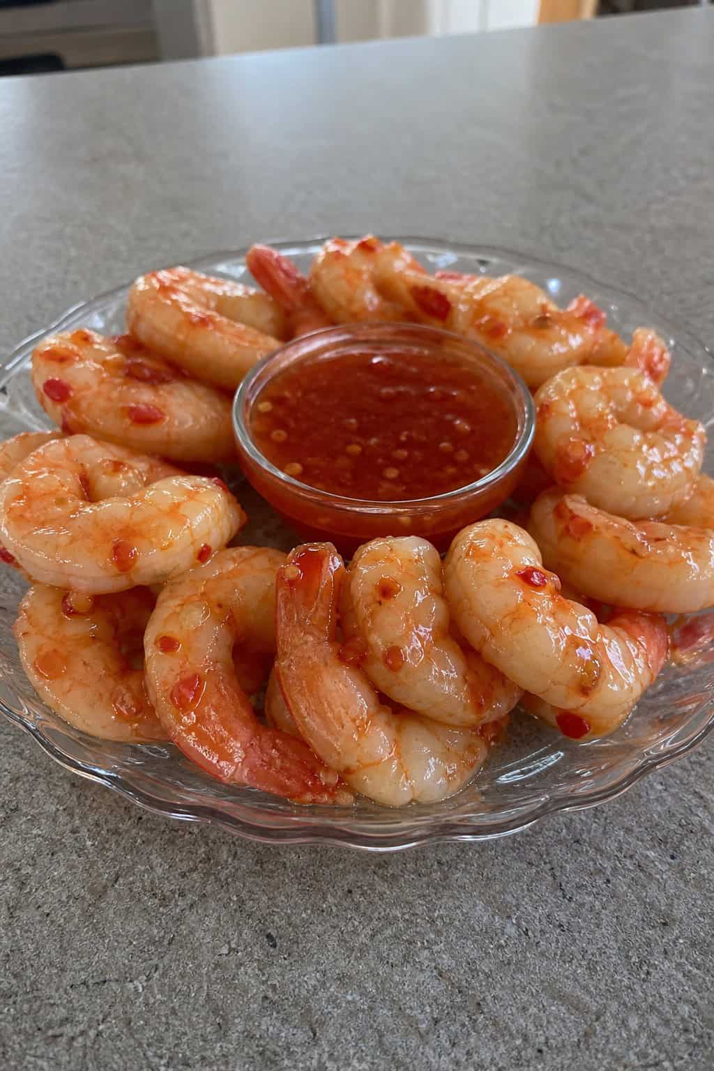A glass dish filled with sweet chili shrimp arranged around a bowl of dipping sauce, garnished with fresh herbs and lime slices on a table.