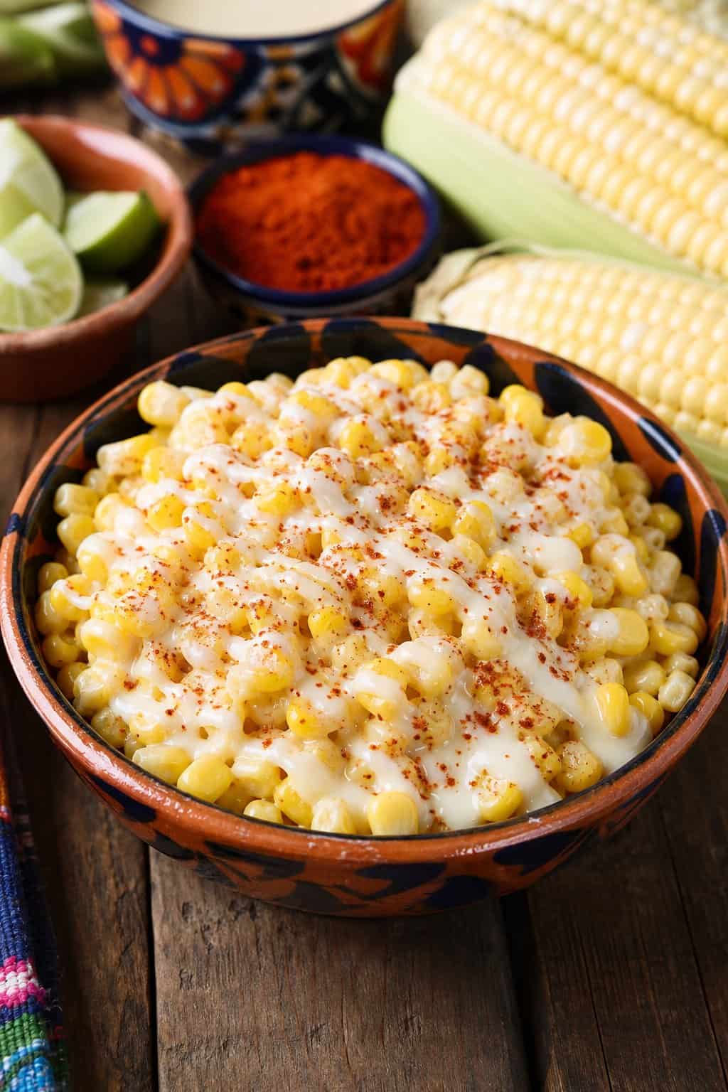 A bowl of sweet corn esquites topped with condensed milk on a wooden table with corn cobs and lime wedges nearby.