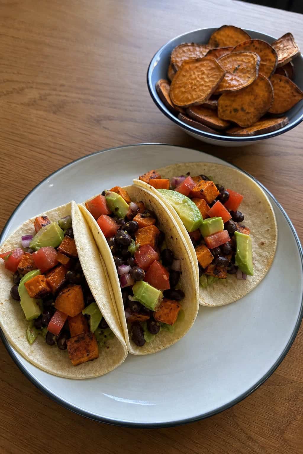 A plate with sweet potato and black bean tacos accompanied by a bowl of roasted sweet potato chips on a table.