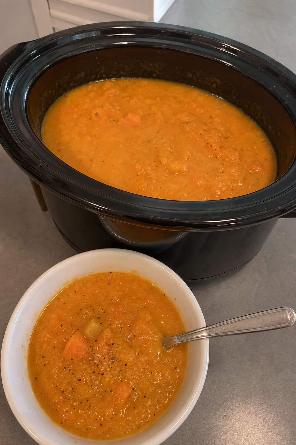 A crockpot filled with steaming sweet potato, apple, and sage soup on a wooden countertop surrounded by fresh sweet potatoes, apples, and sage leaves.