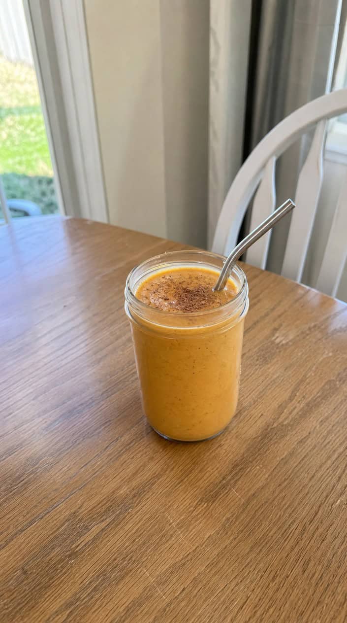 A glass jar of thick orange sweet potato maple smoothie on a wooden table surrounded by two raw sweet potatoes, a small bottle of amber maple syrup, two crossed cinnamon sticks, and a few scattered dried leaves.