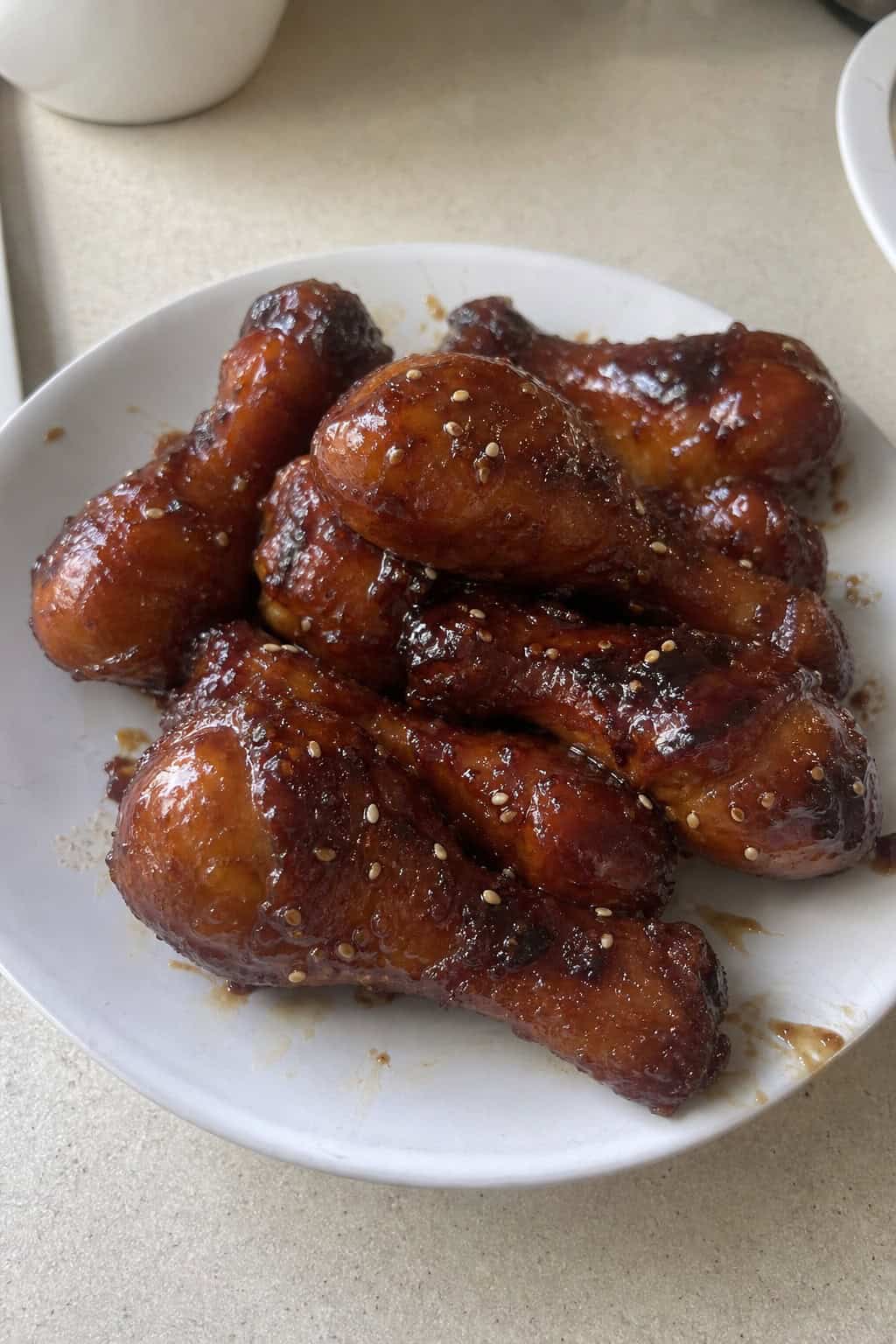 Lacquered sweet soy chicken drumsticks on a dark ceramic plate, finished with white sesame seeds and thinly sliced green onions on a matte wood surface.