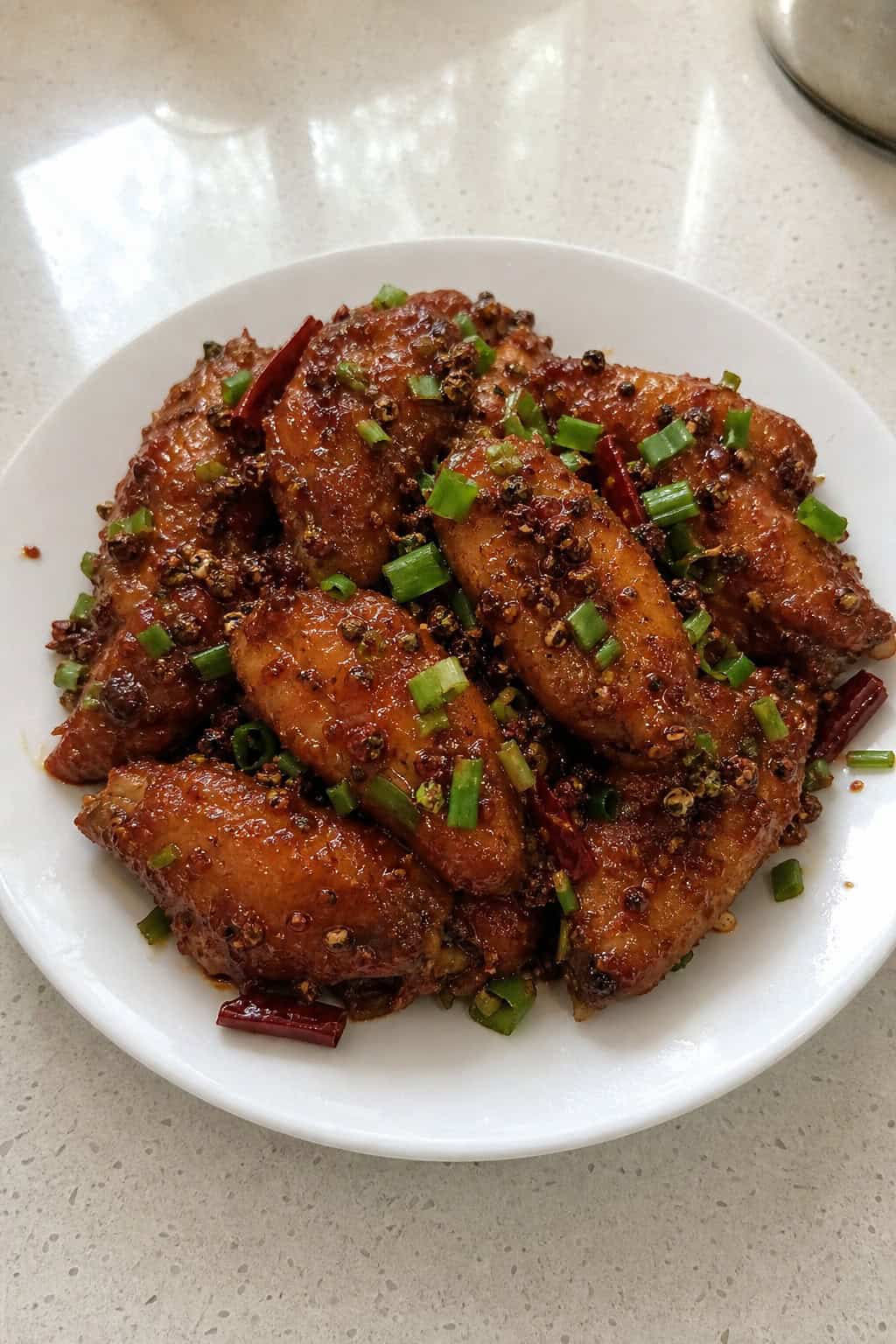 A plate of crispy spicy chicken wings topped with chopped scallions on a wooden table with small bowls of spices and sauce in the background.