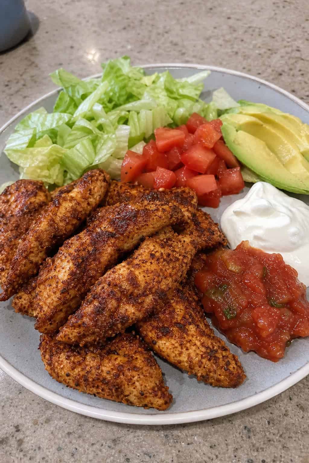 Sliced taco-seasoned chicken tender strips on a plate surrounded by fresh taco toppings including diced tomatoes, sliced avocado, shredded lettuce, cilantro, lime wedges, and small bowls of salsa and sour cream.