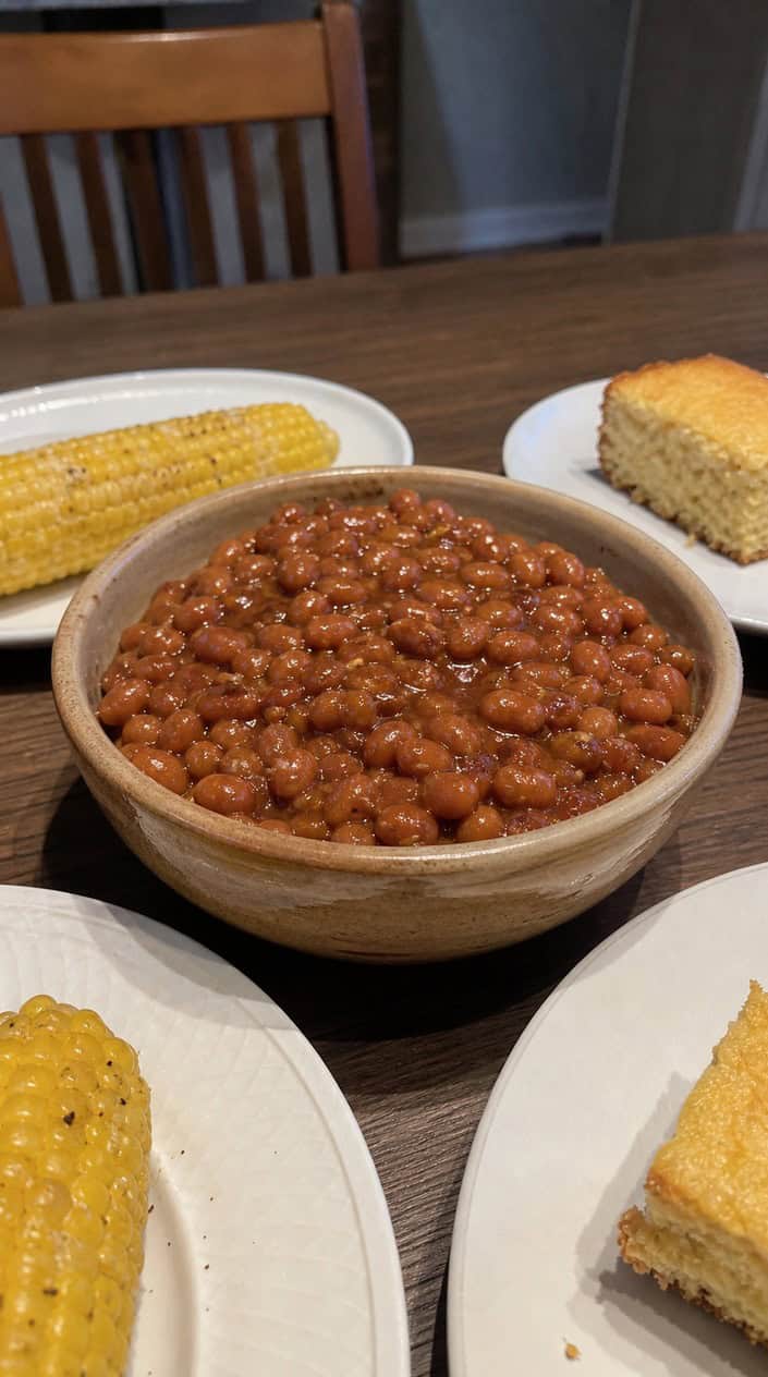 A bowl of baked beans in thick sauce on a wooden table surrounded by BBQ side dishes like corn, coleslaw, and cornbread.