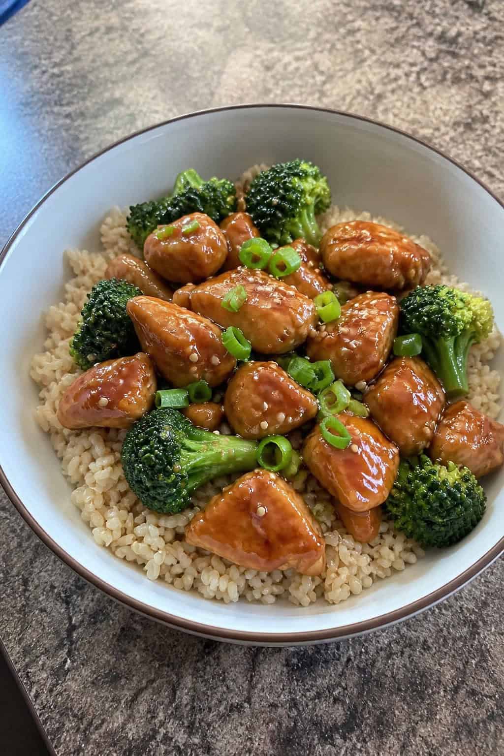 23 Healthy Chicken and Vegetable Recipes 13 A bowl of teriyaki chicken with broccoli and brown rice on a wooden surface.