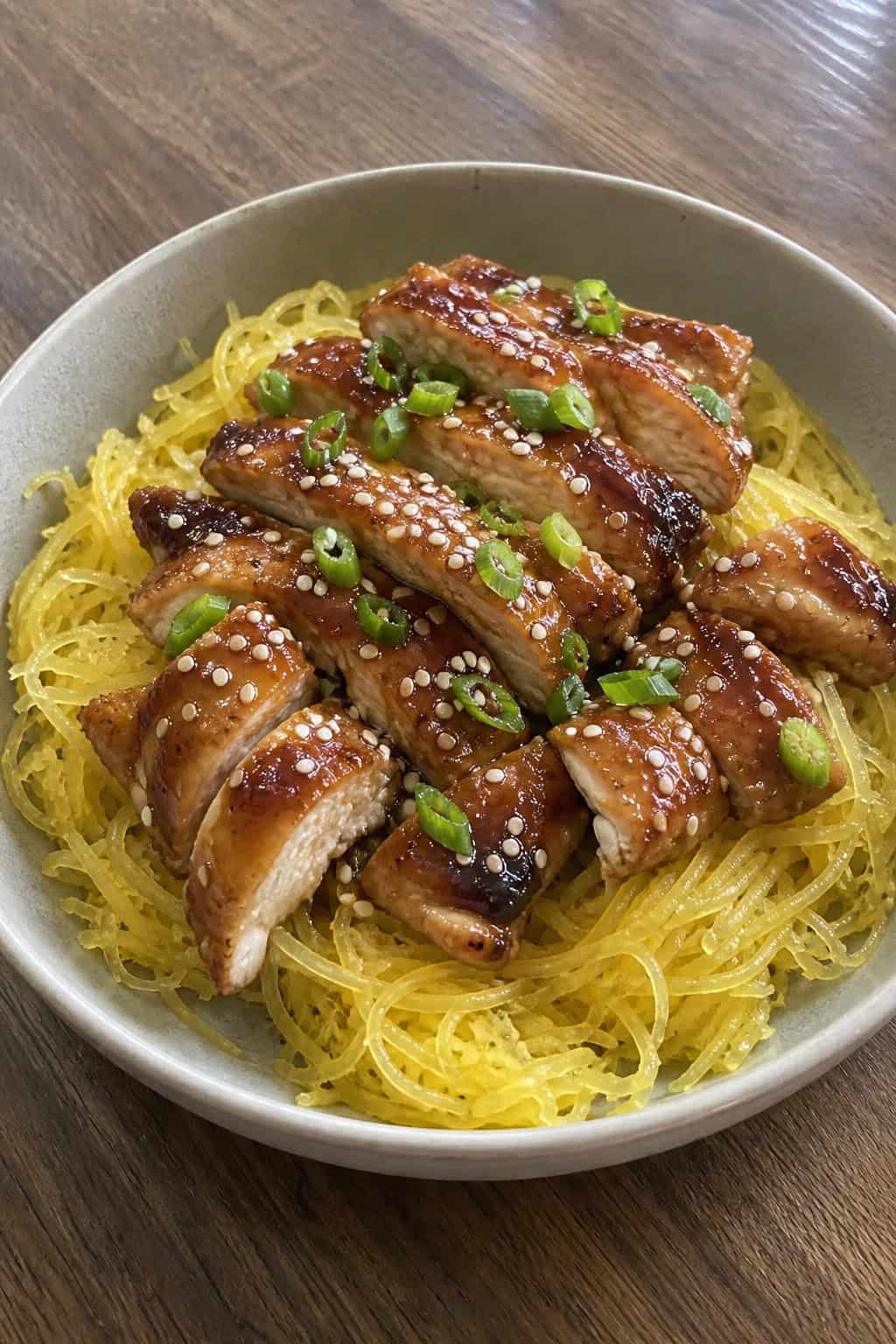 A bowl of teriyaki chicken served over spaghetti squash garnished with green onions and sesame seeds on a wooden table.