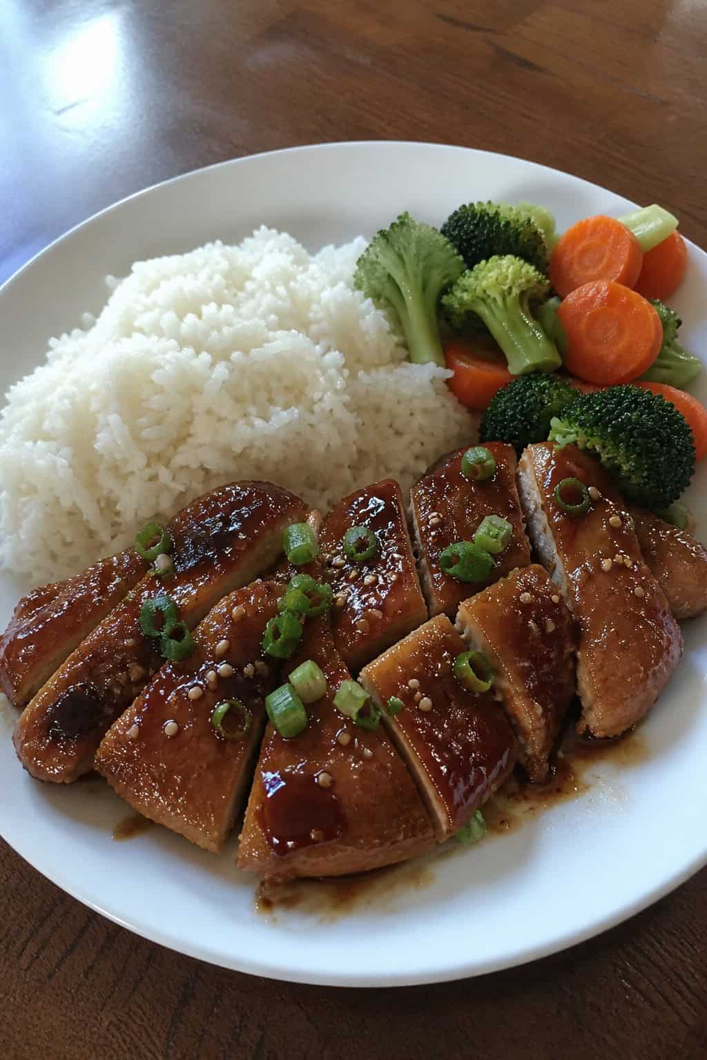 Teriyaki glazed chicken thighs on a white plate with a deep mahogany-colored glaze, served alongside steamed white rice and bright green vegetables.