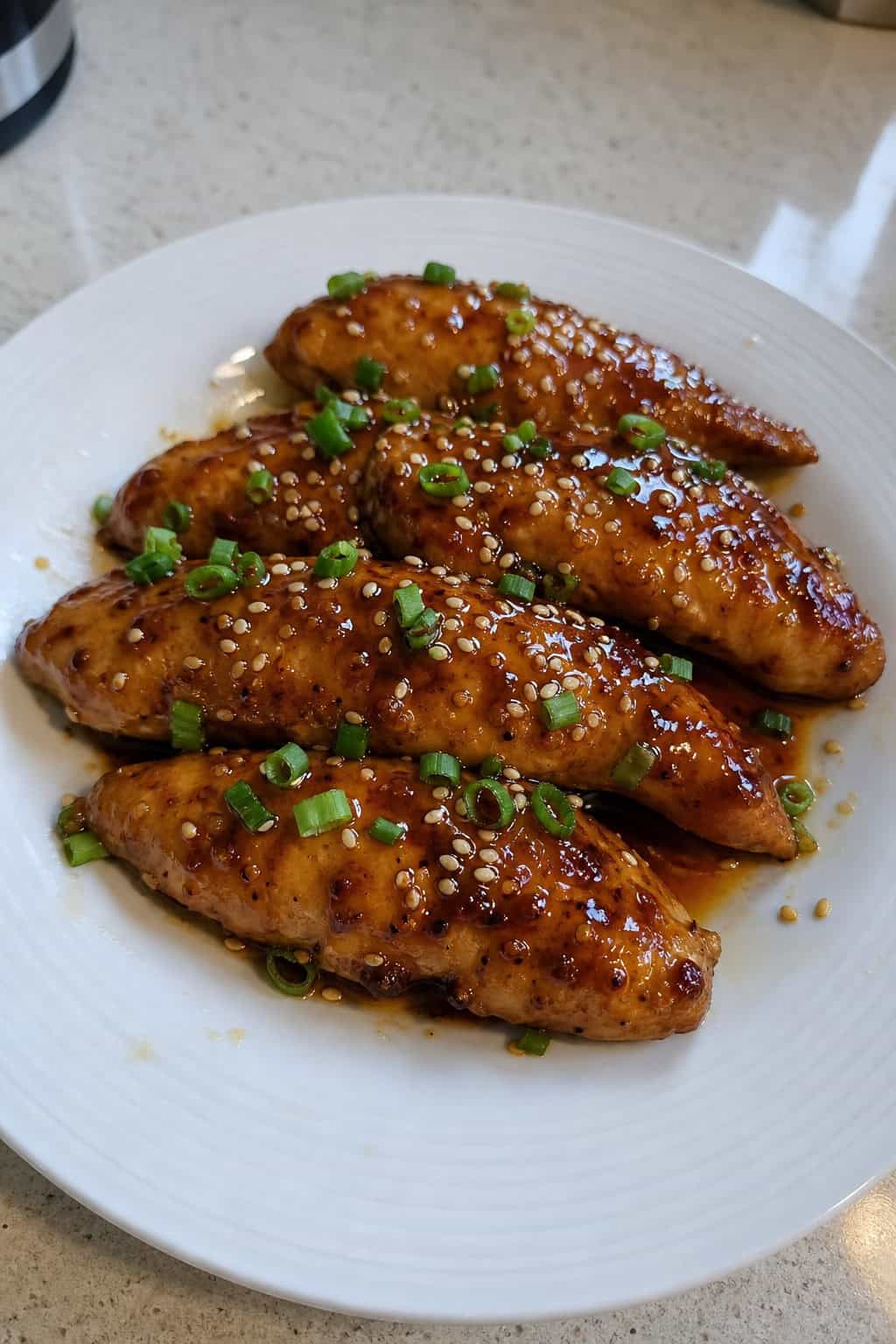 Teriyaki glazed chicken tenderloins topped with sesame seeds and sliced green onions on a white plate over a wooden table.