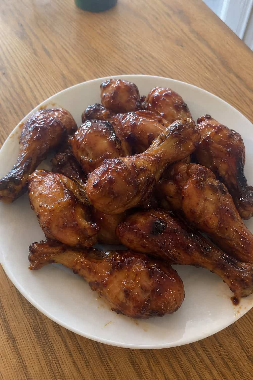 Lacquered teriyaki chicken drumsticks on a light ceramic plate topped with toasted sesame seeds and thinly sliced green onions, with small sauce bowls and fresh ginger visible at the edges of a wooden table.