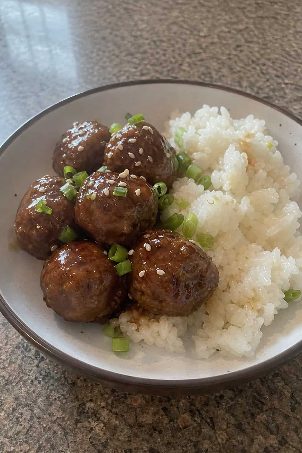 A bowl of teriyaki meatballs with rice, garnished with green onions and sesame seeds, on a wooden table with fresh ingredients nearby.