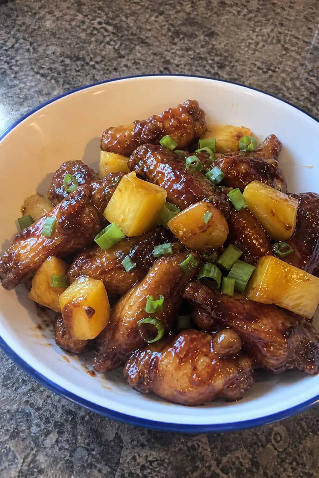 A plate of glazed teriyaki chicken wings with pineapple chunks and green onion garnish on a wooden table.