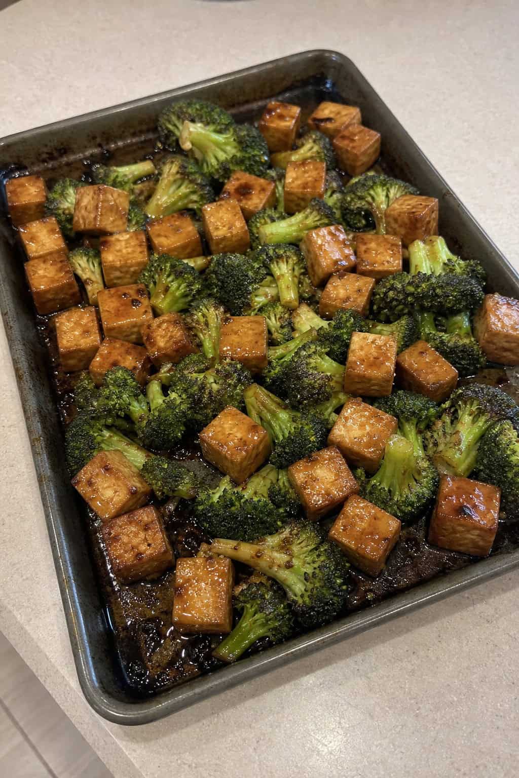 Golden-edged cubed firm tofu and deep-charred broccoli florets coated in a dark teriyaki glaze on a foil-lined sheet pan on a wooden table, garnished with sesame seeds and sliced green onions.