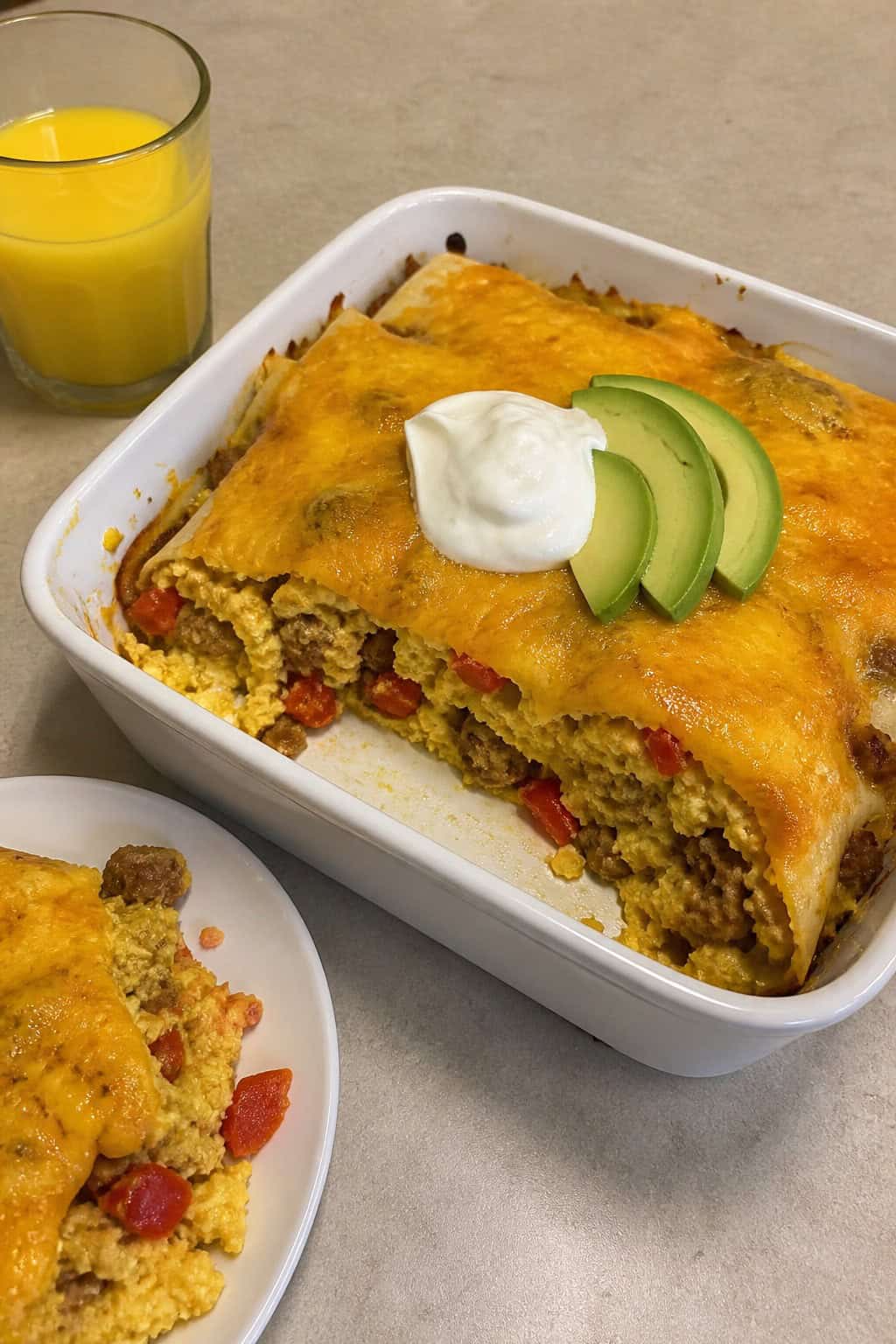 A baked Tex-Mex breakfast enchilada casserole topped with golden melted cheese, sliced avocado, and fresh cilantro in a rectangular baking dish, with a small glass of orange juice and a bowl of salsa on the table.