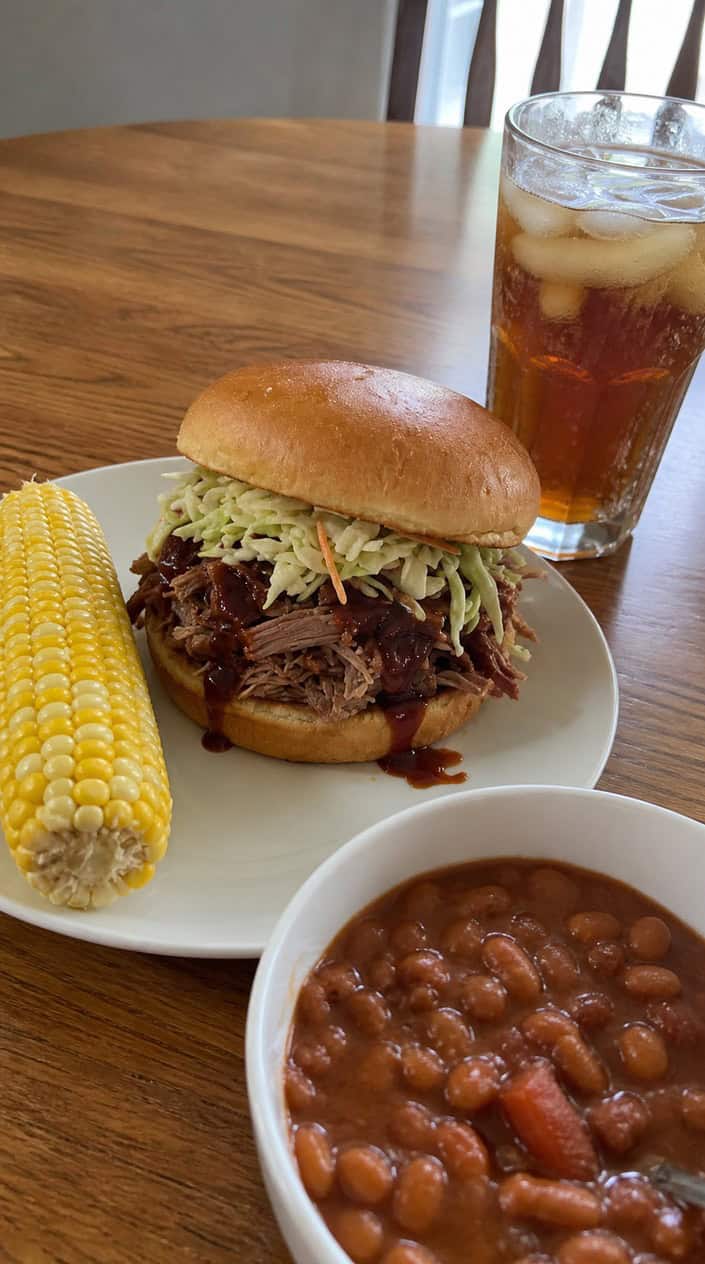A pulled pork sandwich with barbecue sauce and coleslaw on a bun, placed on a wooden table with summer picnic foods and people celebrating in the background.