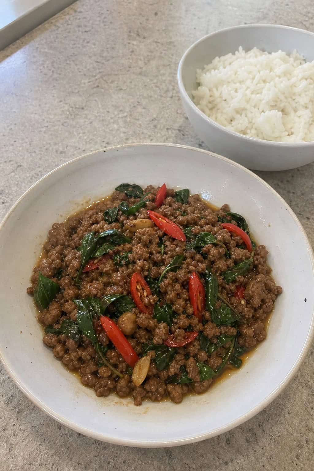 A bowl of Thai Basil Beef with ground beef, green basil leaves, and red chili peppers, served with steamed rice on a wooden table.