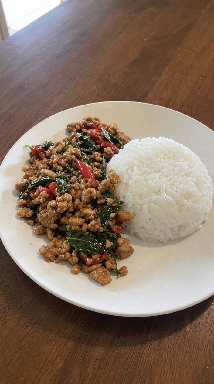 A plate of Thai basil ground chicken with jasmine rice on a wooden table, garnished with fresh basil and red chili slices.
