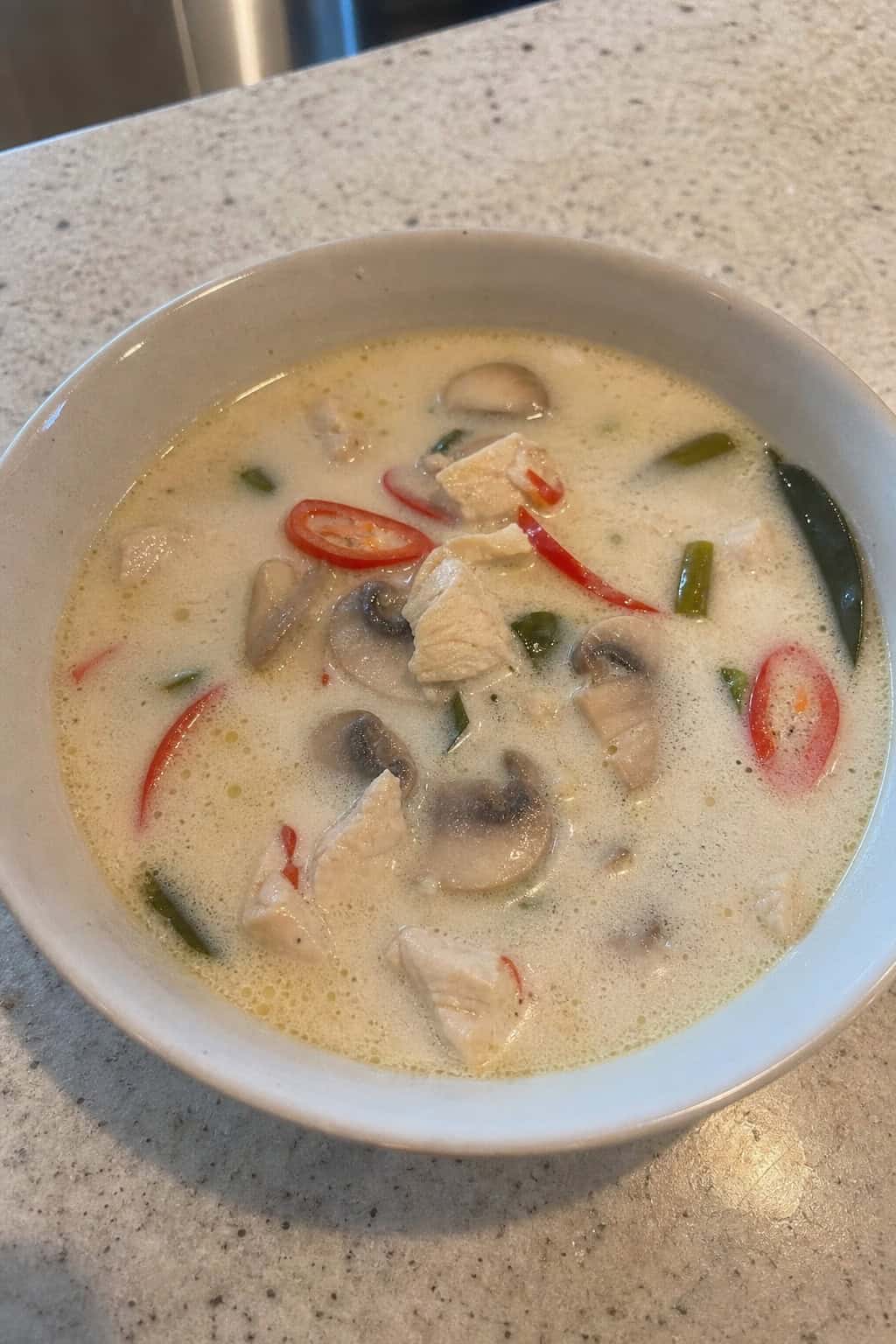 A bowl of Thai coconut chicken soup with chicken, mushrooms, cilantro, and chili on a wooden table surrounded by fresh herbs and ingredients.