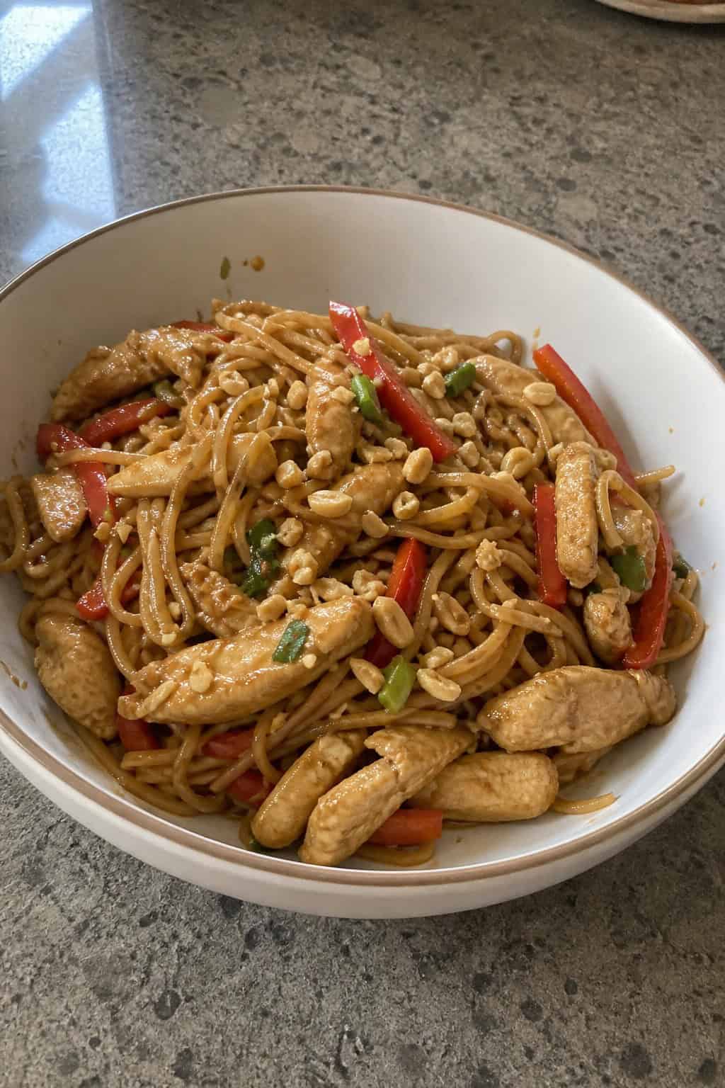 A bowl of Thai peanut chicken and noodles garnished with fresh cilantro and crushed peanuts, with chopsticks resting on the bowl on a wooden table.