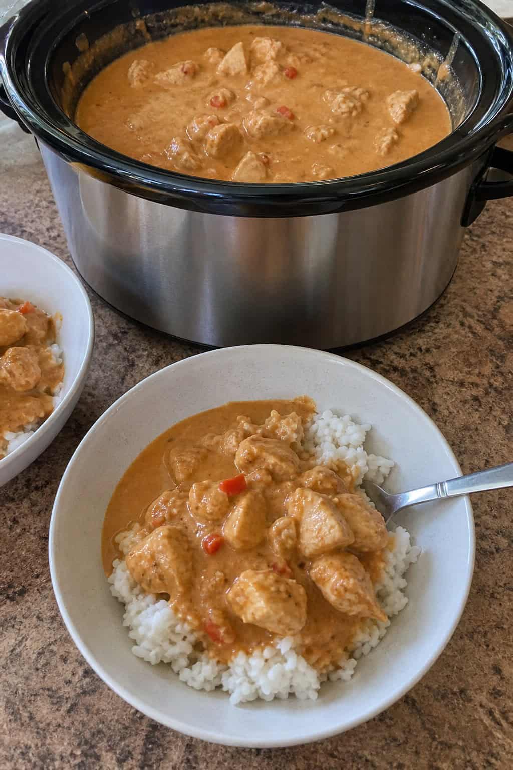 A slow cooker filled with Thai peanut chicken surrounded by fresh ingredients on a wooden table.