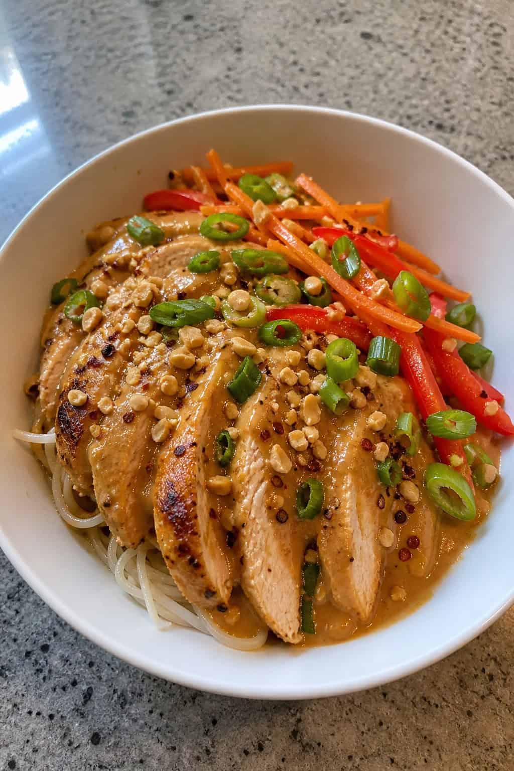 A bowl of Thai peanut chicken noodle dish with grilled chicken, noodles, fresh vegetables, crushed peanuts, and a lime wedge.