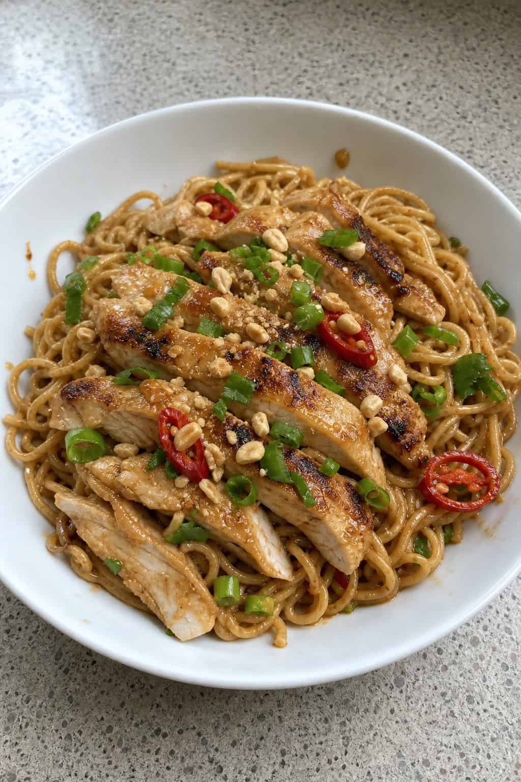 A bowl of Thai peanut chicken noodles garnished with green onions, crushed peanuts, cilantro, and red chili slices on a wooden table.