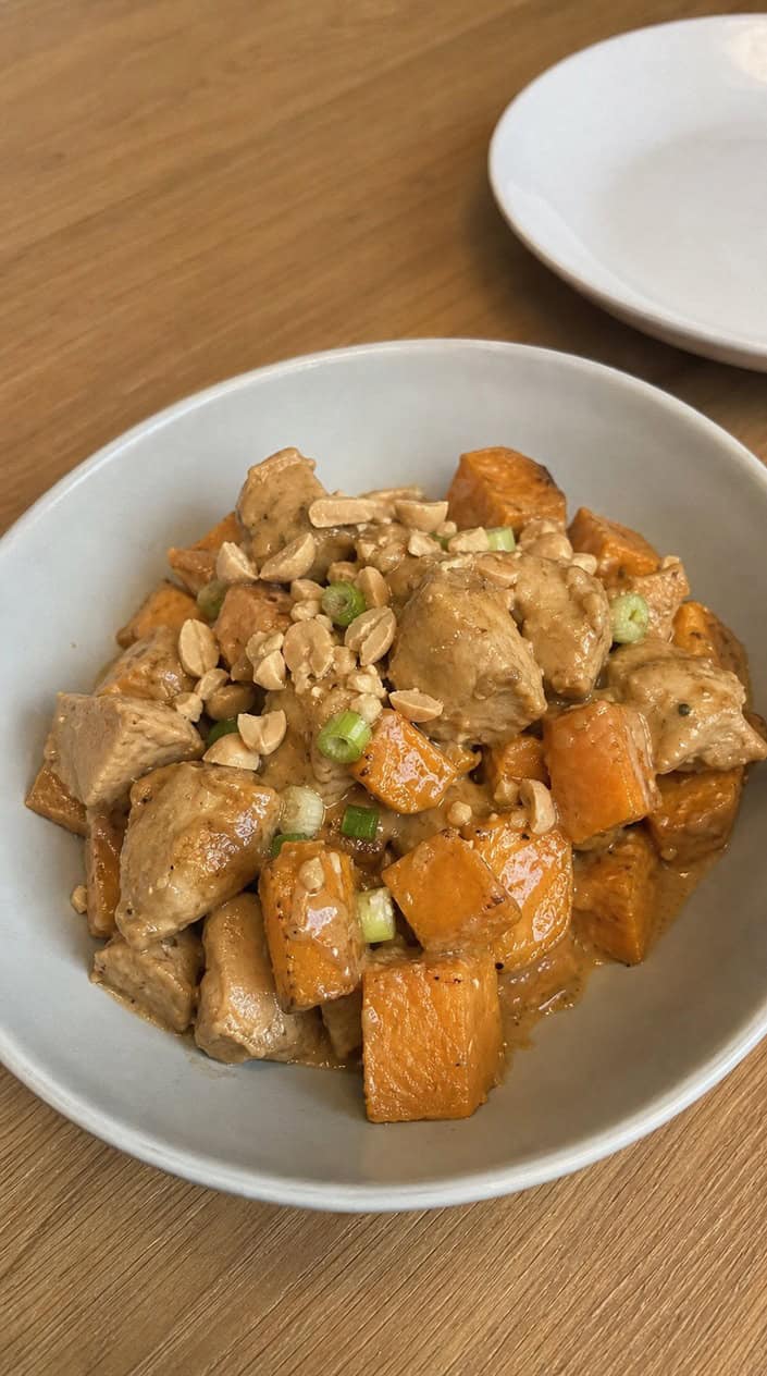 Bowl of Thai peanut chicken with roasted sweet potatoes garnished with cilantro and peanuts on a wooden table.