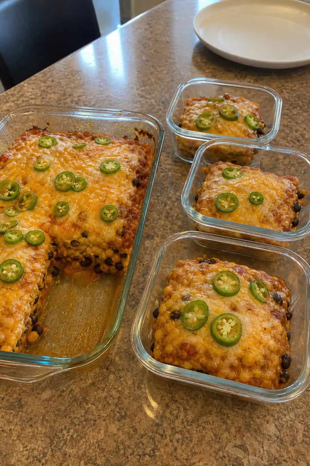 A kitchen countertop with a partially sliced baked enchilada casserole beside a row of clear glass storage containers being filled with individual portions.