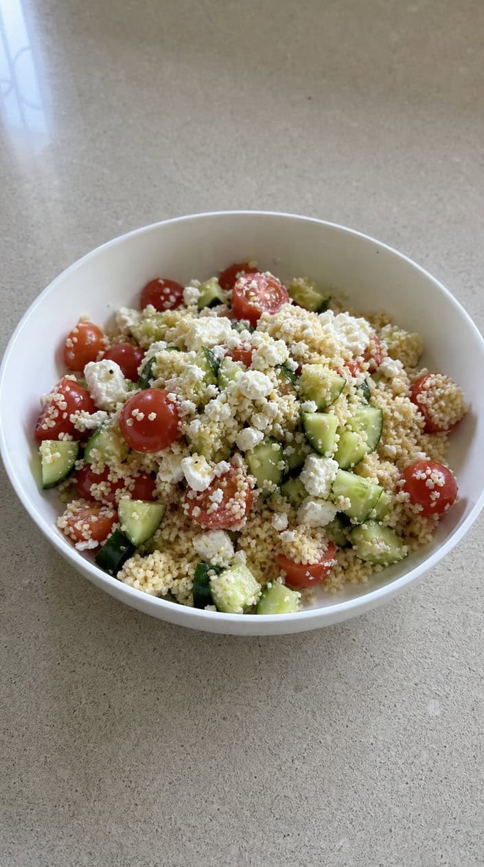 A bowl of tomato, cucumber, and feta couscous salad on a wooden table with fresh herbs and a glass of iced water nearby.