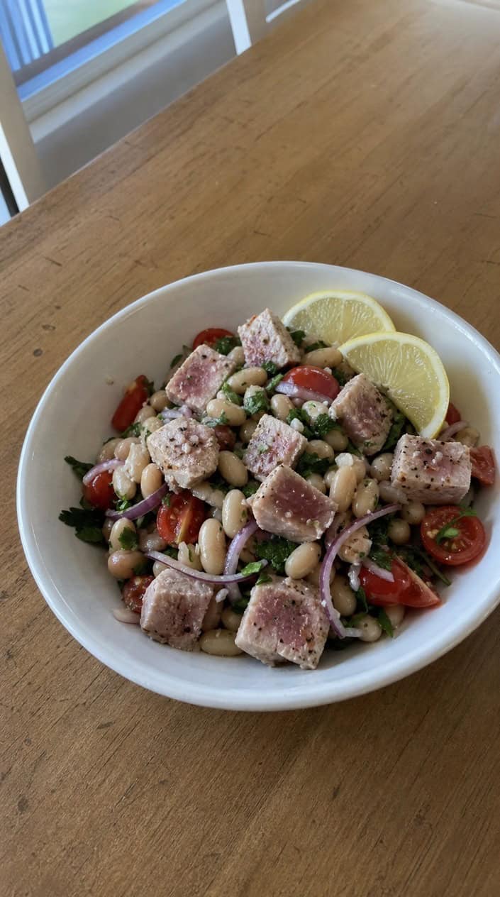 A bowl of tuna and white bean salad garnished with lemon slices and parsley on a wooden table.