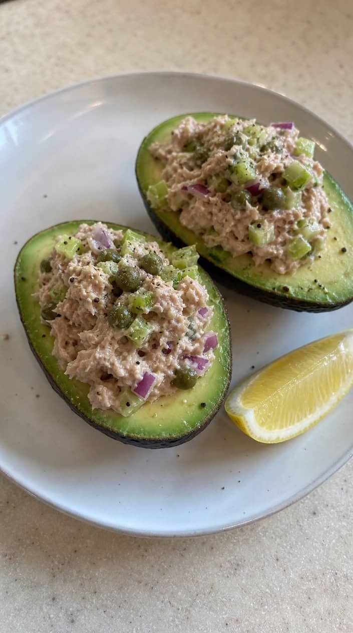 Two avocado halves stuffed with tuna salad containing celery and capers, placed on a wooden board with fresh herbs and lemon wedges in the background.