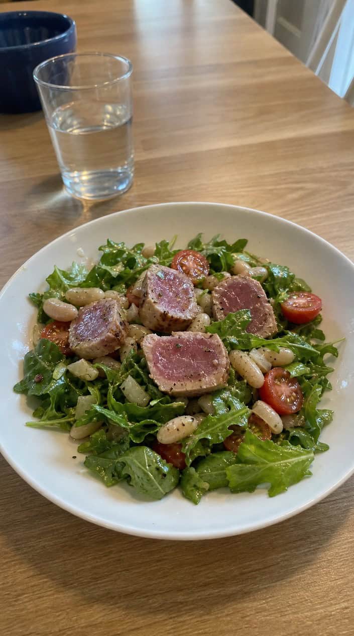 A bowl of tuna, white bean, and arugula salad with lemon vinaigrette on a wooden table, accompanied by a lemon wedge and a small jar of dressing.