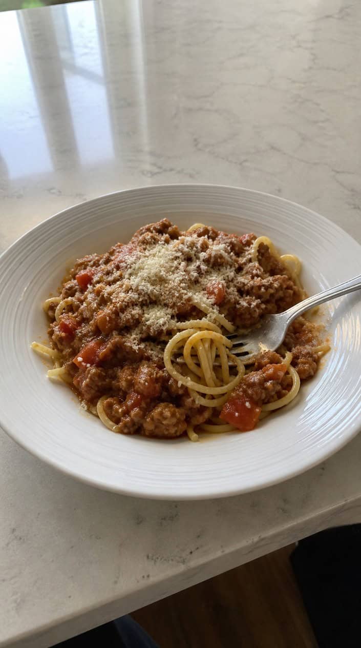 A bowl of turkey Bolognese sauce served over pasta or zucchini noodles on a wooden table with fresh ingredients around it.
